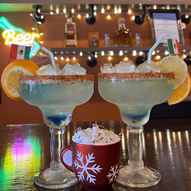 Two blue margaritas with orange and lime slices, and a red snowflake mug on a bar.
