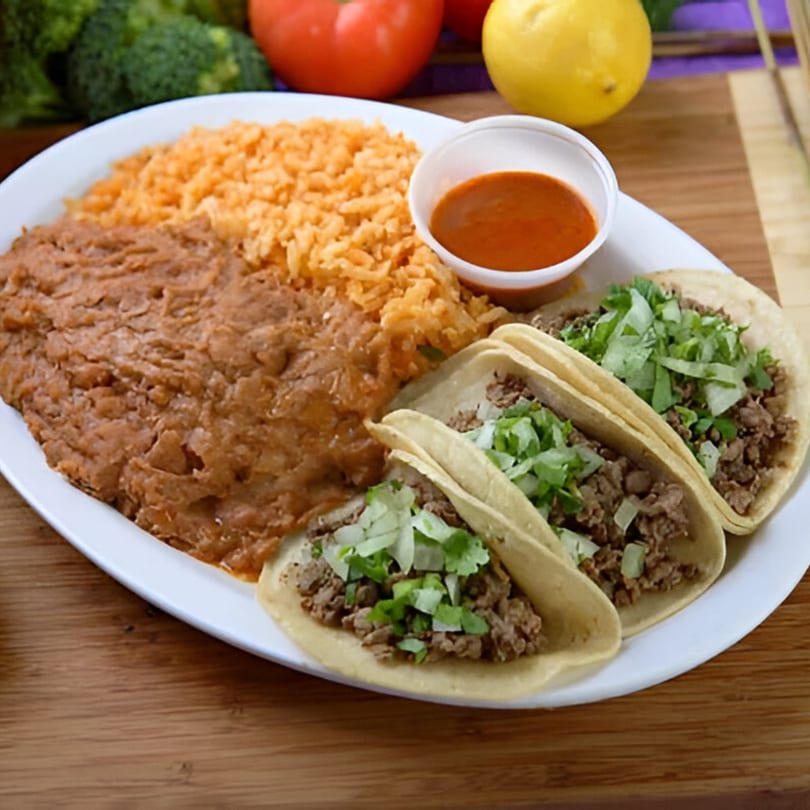 A plate of three carne asada tacos with rice, refried beans, and a side of red sauce.