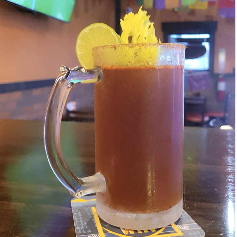 A frosty mug of Michelada, garnished with a lime wedge and celery, on a wooden bar.