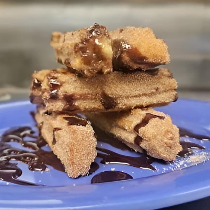 Golden brown churros, drizzled with chocolate sauce, stacked on a vibrant blue plate.