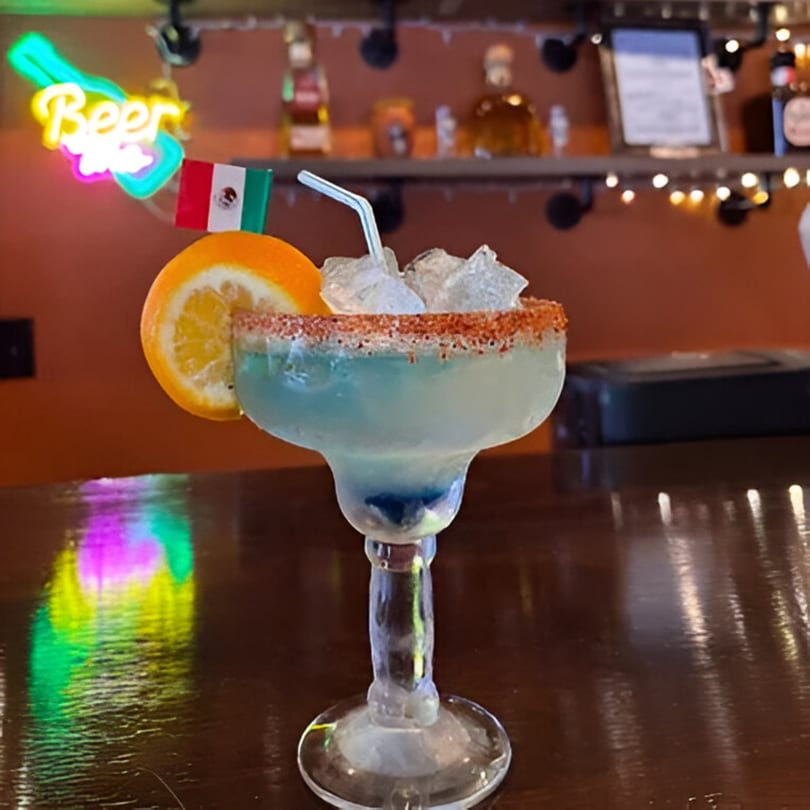 A blue margarita with a salted rim, orange slice, and Mexican flag on a bar counter.