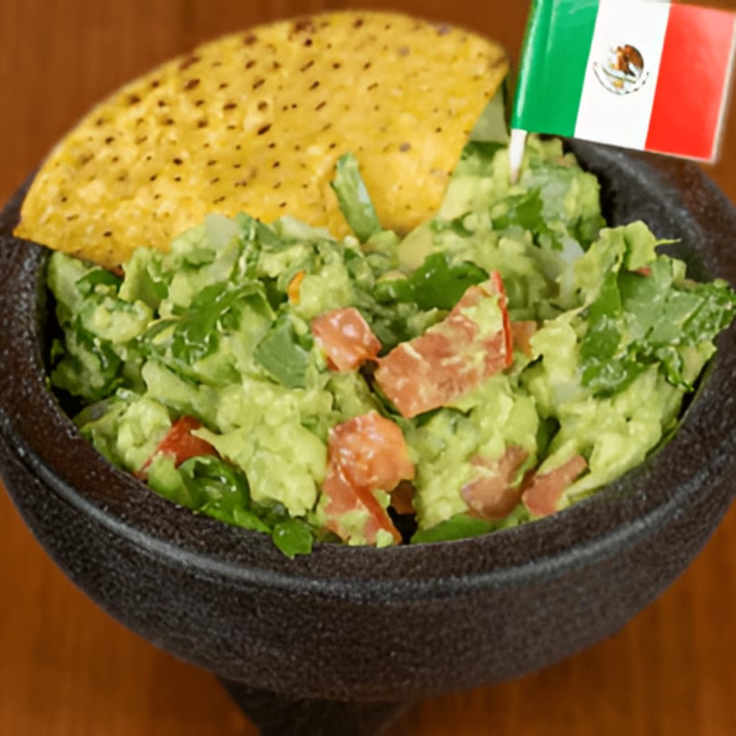 Fresh guacamole with diced tomatoes and cilantro, a tortilla chip, and a Mexican flag in a dark bowl.