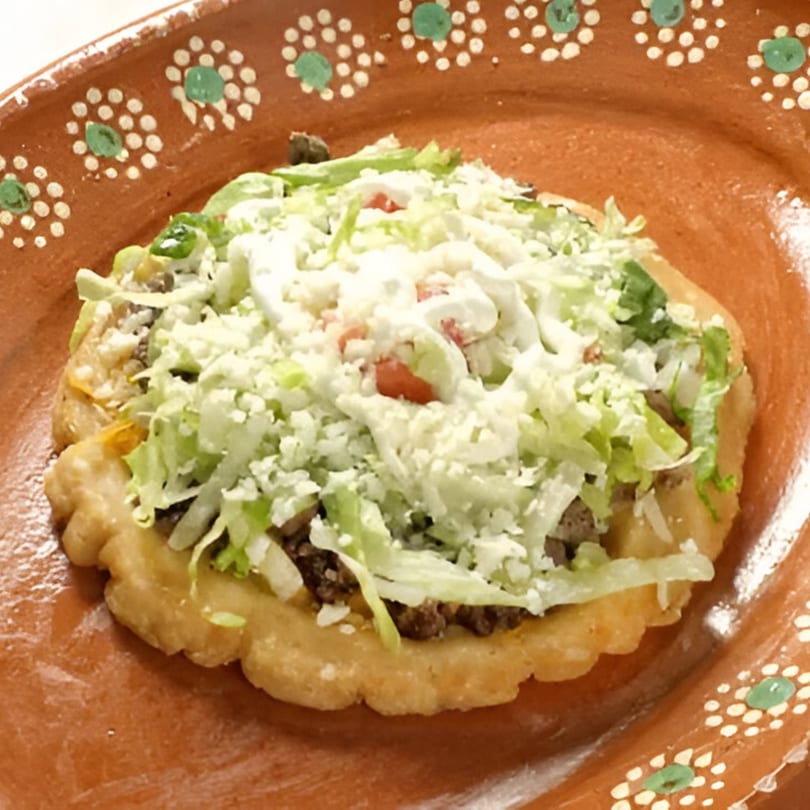 A Taco Station sopes with ground beef, lettuce, cheese, and sour cream on a decorative clay plate.