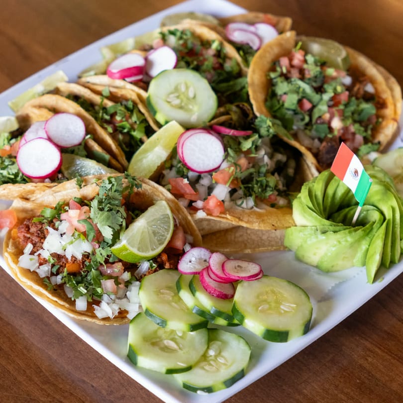 A platter of Taco Station tacos with various toppings, sliced radishes, cucumbers, and fanned avocado.